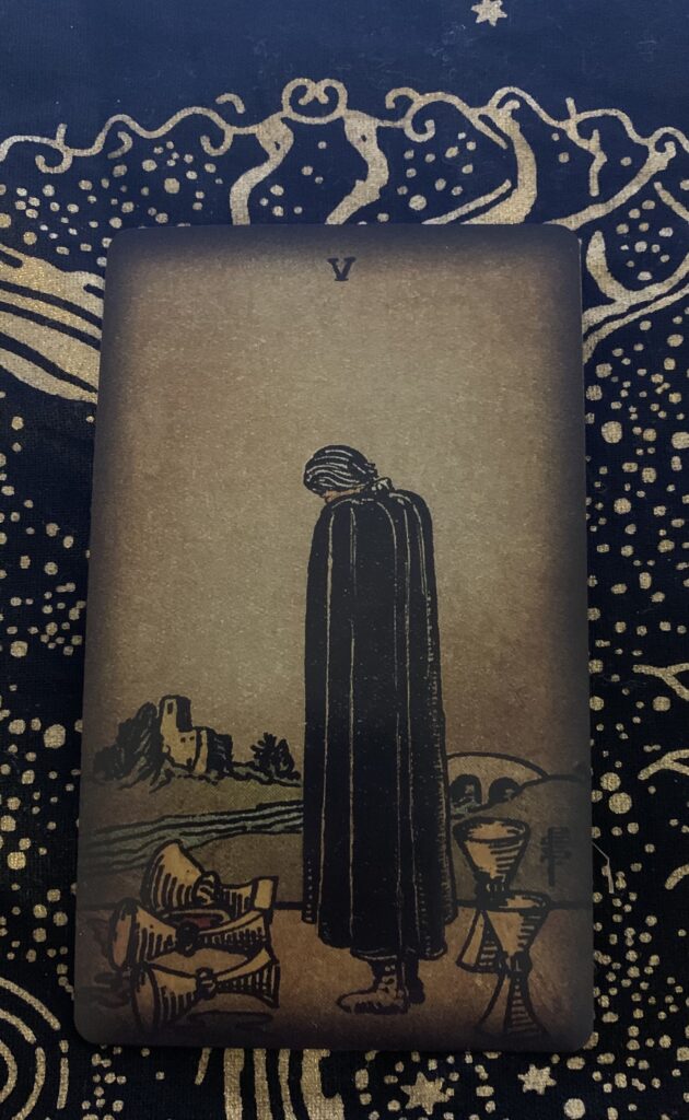 The 5 of Cups depicts a man wearing a black cape with three overturned cups in front of him with two upright behind him. A river flows under an arched bridge with a small castle on the other side.